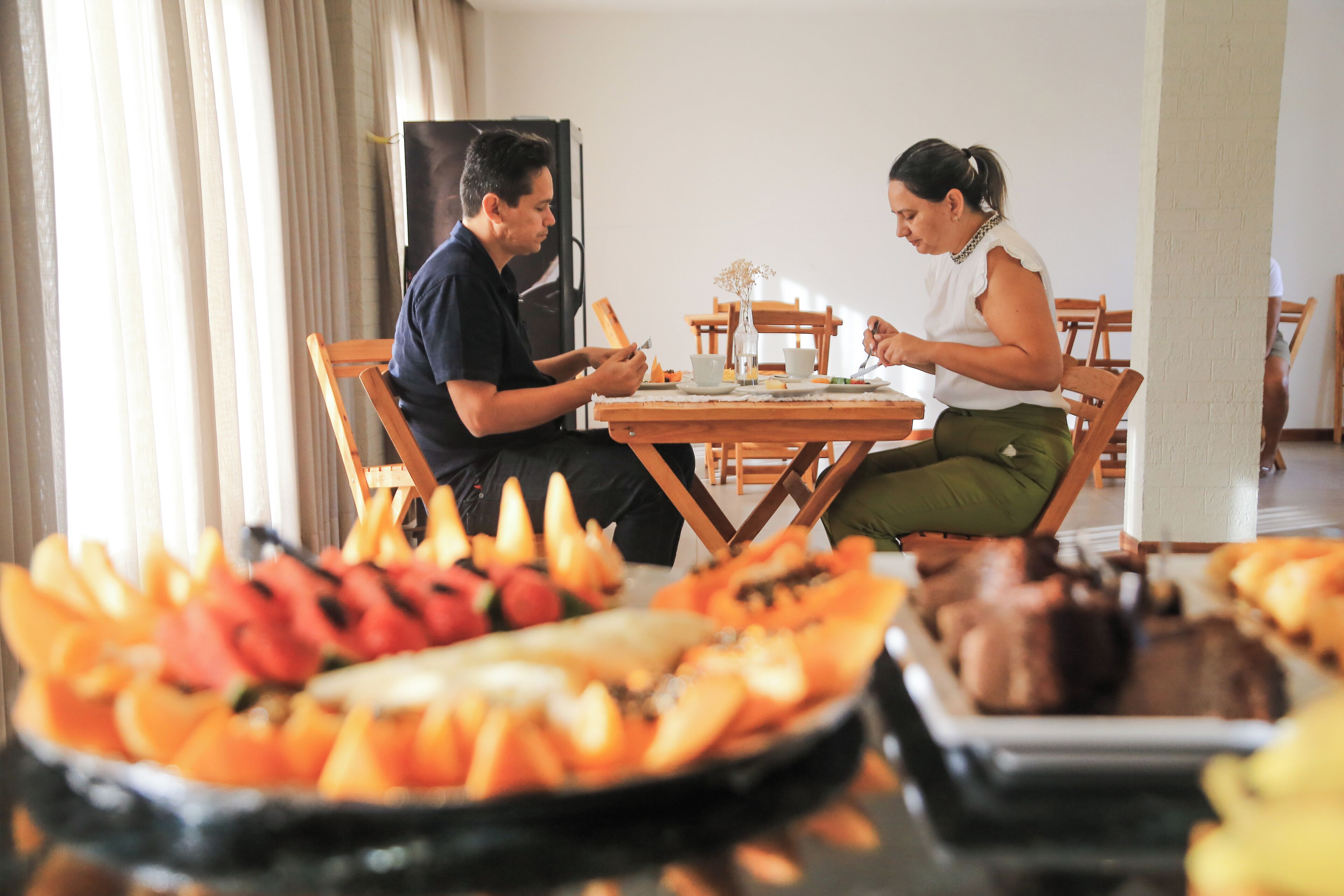 A dining area with a table set for two, surrounded by various fruits and a meat dish.