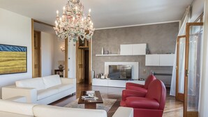 Een moderne woonkamer met een witte bank, een rode fauteuil, een kroonluchter en een keukenhoek met witte kasten en een televisie.
