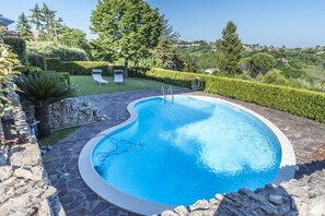 Una piscina con borde de piedra, dos sillas reclinables y vista a un paisaje verde.