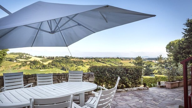 Een terras buiten met een tafel, stoelen, een grote parasol en een prachtig uitzicht over glooiende heuvels.