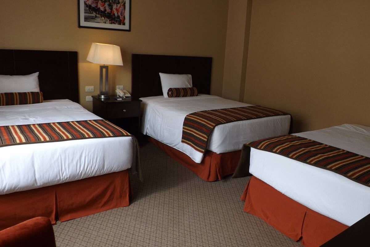 A hotel room with three single beds, each with a striped bedspread and white pillows. There is a nightstand with a lamp and a telephone between the beds.