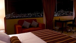 A hotel room with a bed, a red armchair, a desk, and a view of a cityscape at night. - Apu Mountain Lodge (Cusco)