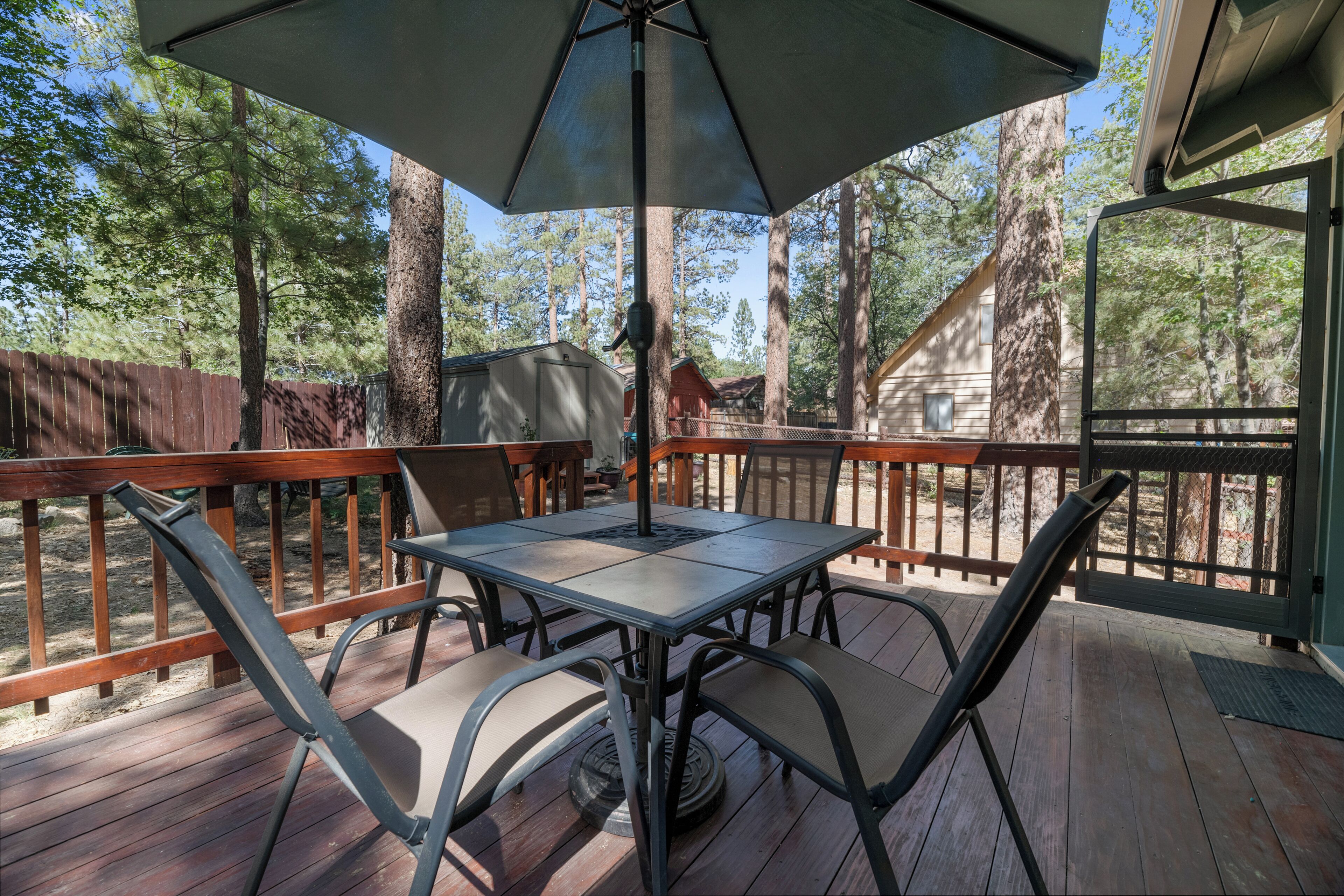 A covered outdoor deck with a table and chairs, surrounded by a wooden deck and tall trees.