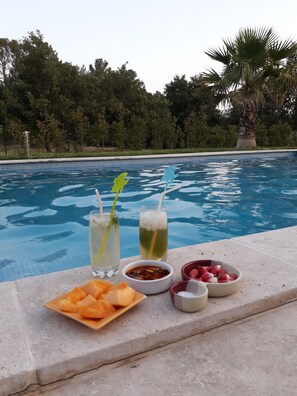 A poolside table with a plate of fruit, two drinks, and small bowls of condiments.