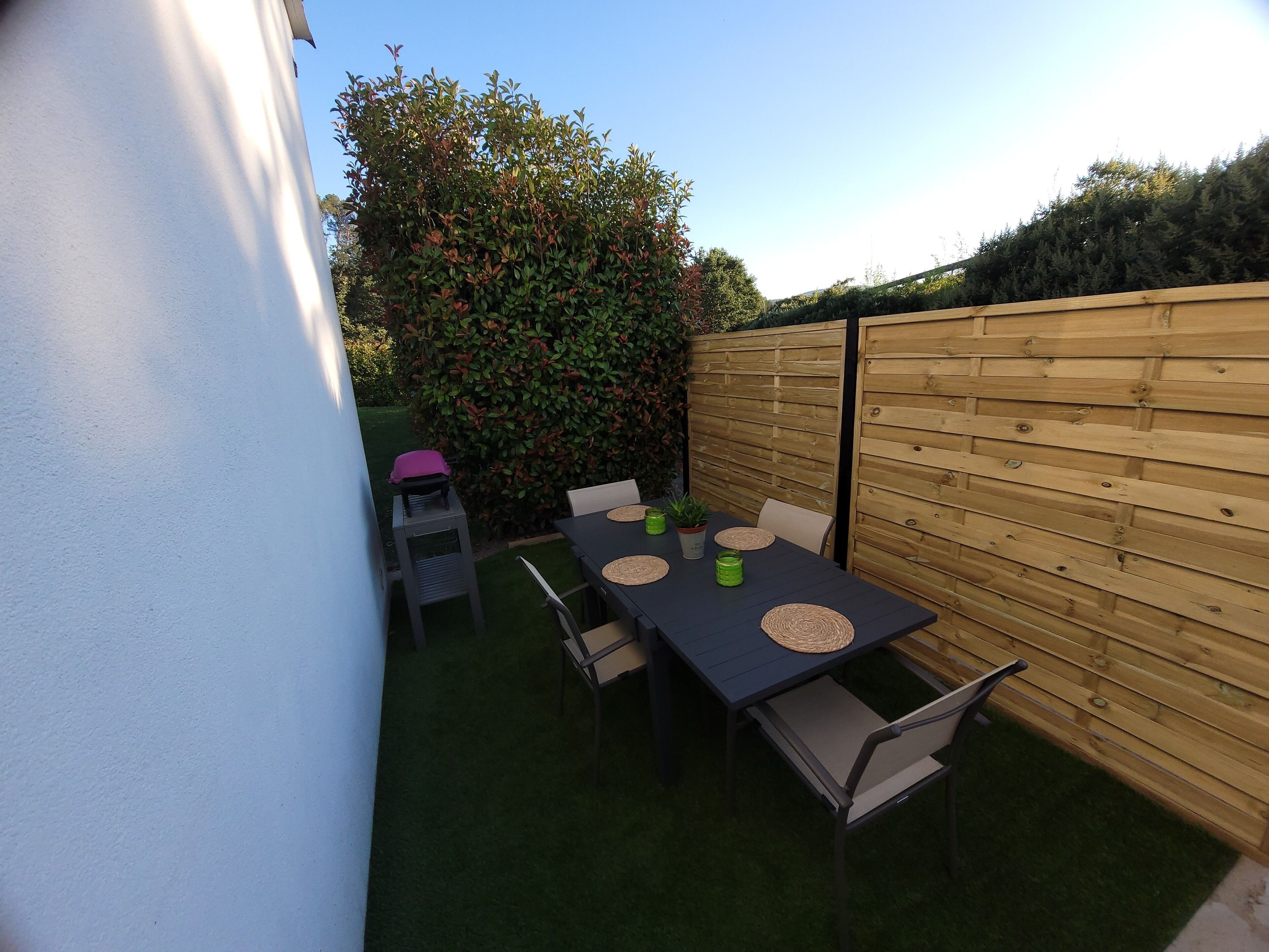 Eine Terrasse mit Tisch und Stühlen, umgeben von einem Holzzaun und einem Heckenstreifen.