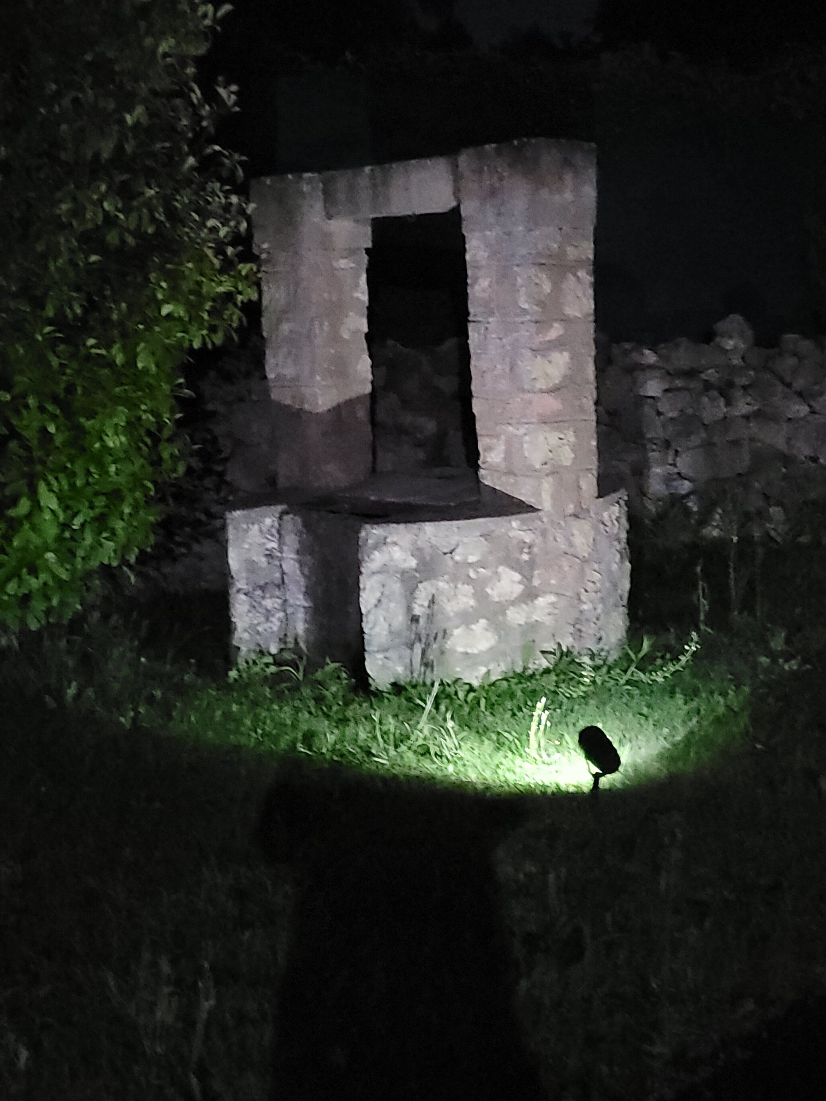A stone structure with a rectangular opening, surrounded by greenery and a small bird on the grass.