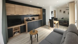A modern living room with a grey sofa, a wooden coffee table, and a kitchen area with wooden cabinets and a black countertop.