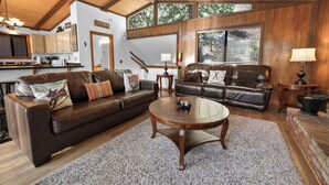 A spacious living room with two leather sofas, a wooden coffee table, and a kitchen in the background. - Treetop Falls (Big Bear Lake)