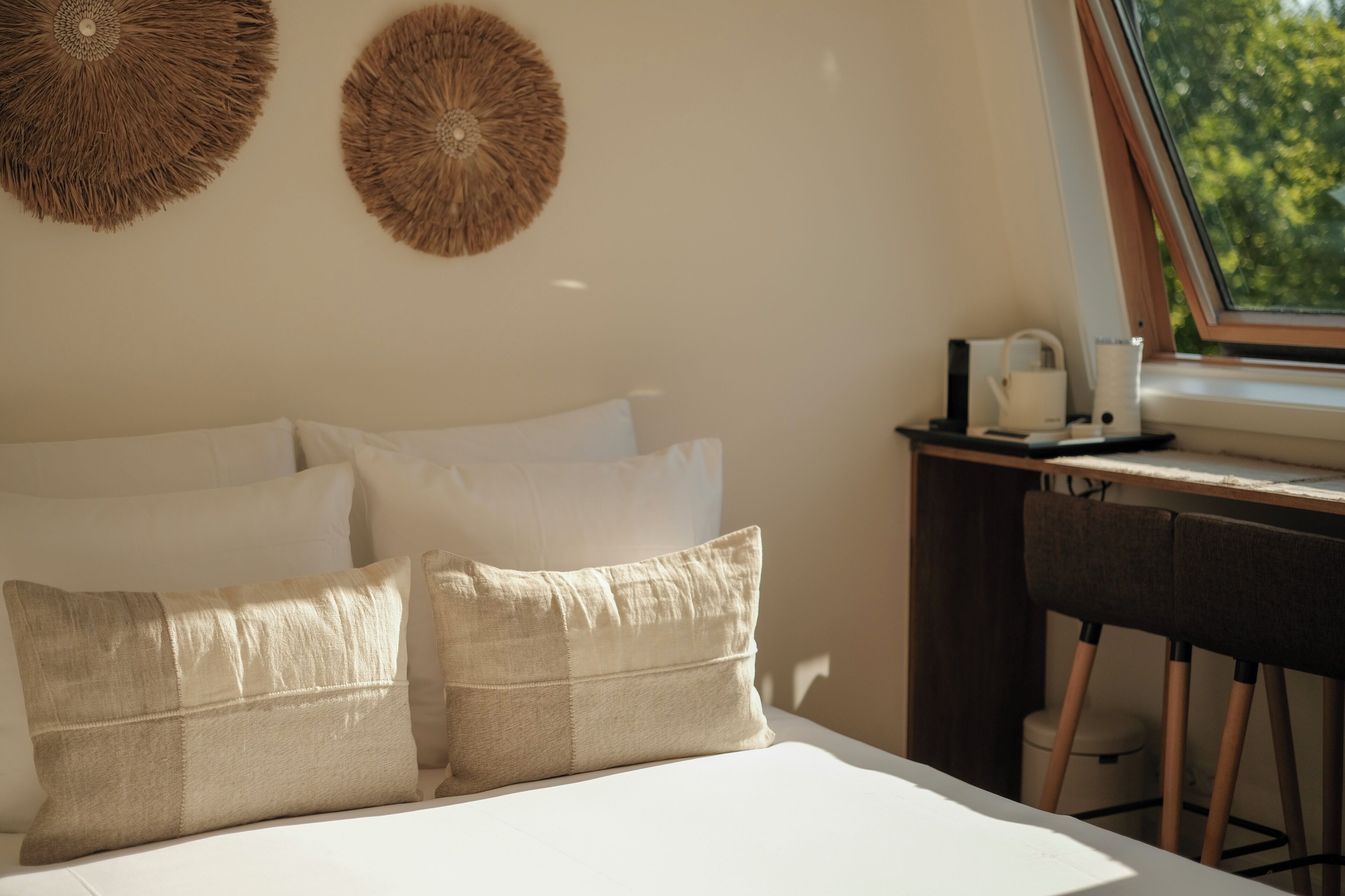 A bed with white linens and beige pillows, a small table with a kettle, and a window with a view of trees.