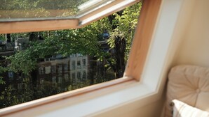 A room with a window overlooking a canal, a chair, and a view of buildings and trees. - Canal View Stay Amsterdam (Amsterdam)