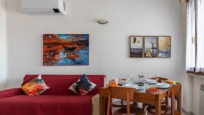 Ein Wohnzimmer mit einer roten Couch, einem Esstisch gedeckt mit Tellern, Gläsern und einer Obstschale. An der Wand hängen drei gerahmte Bilder.