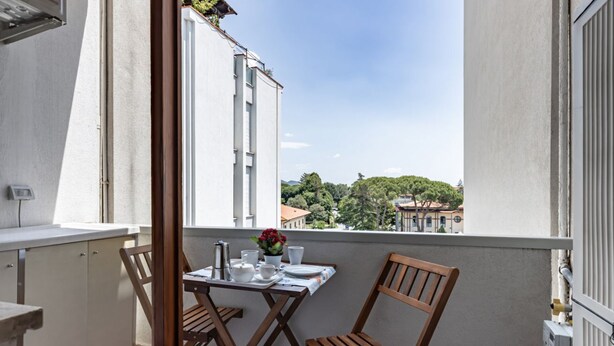 Ein Balkon mit einem Tisch für zwei Personen, zwei Holzstühlen und Blick auf Gebäude und Bäume im Hintergrund.