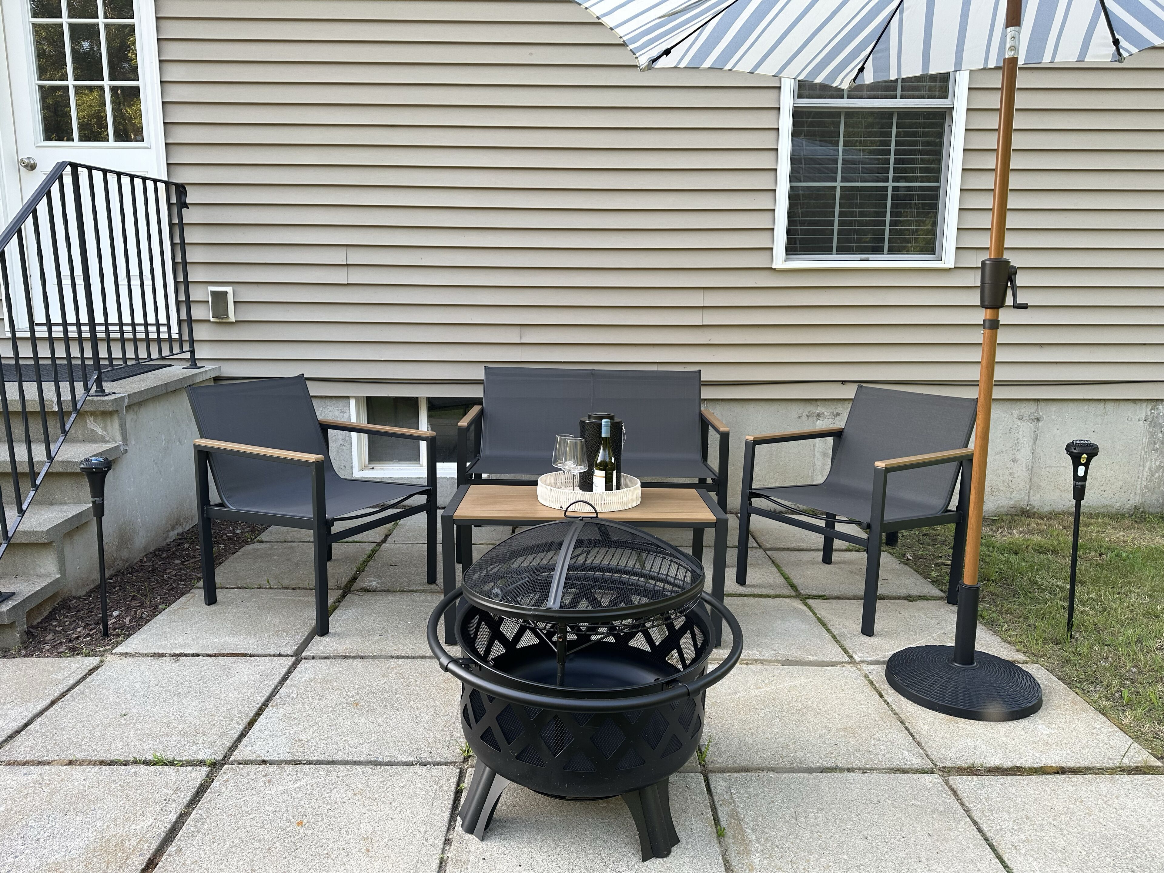 Une terrasse extérieure avec un foyer en métal noir, deux chaises, une petite table et un parasol à rayures.