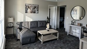 A modern living room with a sectional sofa, a coffee table, a lamp, a painting of a yak, a ceiling fan, and a mirror. - The Hidden Oasis, Duplex A. Close to almost everything! (Enfield)