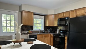 A modern kitchen with wooden cabinets, a black refrigerator, and a dining area with a table set for four. - The Hidden Oasis, Duplex A. Close to almost everything! (Enfield)
