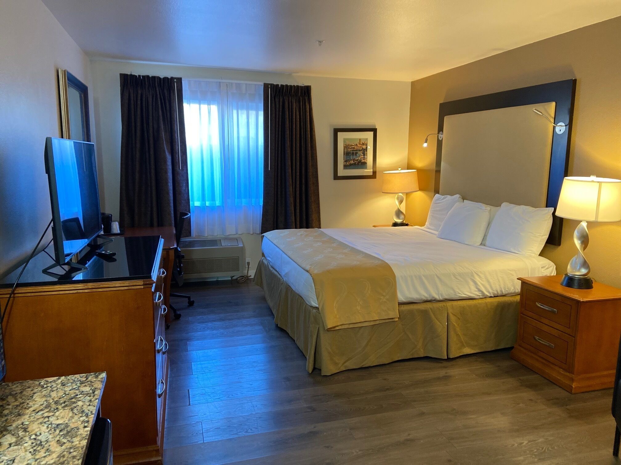 A hotel room with a bed, a desk with a computer, a television, a window with curtains, and a wall-mounted lamp.