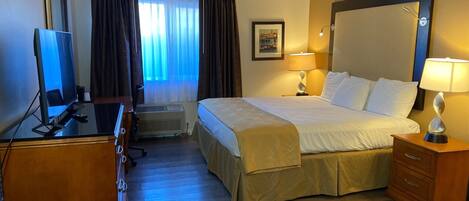 A hotel room with a bed, a desk with a computer, a television, a window with curtains, and a wall-mounted lamp.