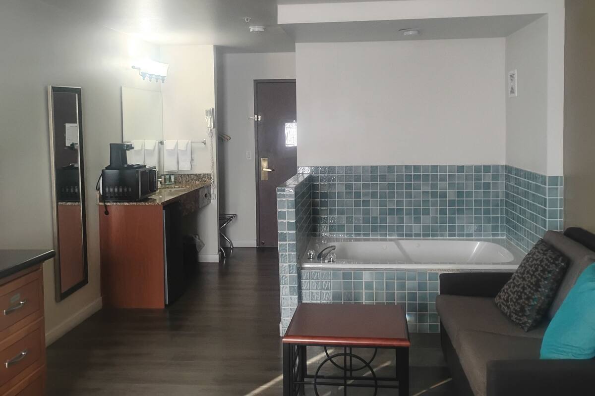 A hotel room with a bed, a sofa, a small table, a microwave, a dresser, a mirror, and a bathroom with a bathtub and tiled walls.