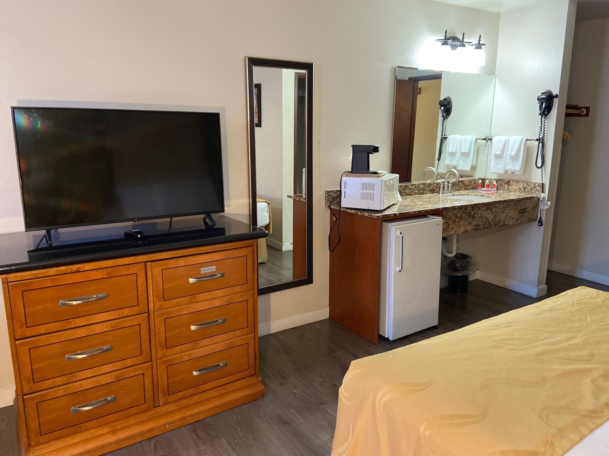 Habitación de hotel con tocador de madera, televisor, refrigerador pequeño, lavamanos con espejo y cama con cubrecama amarillo.
