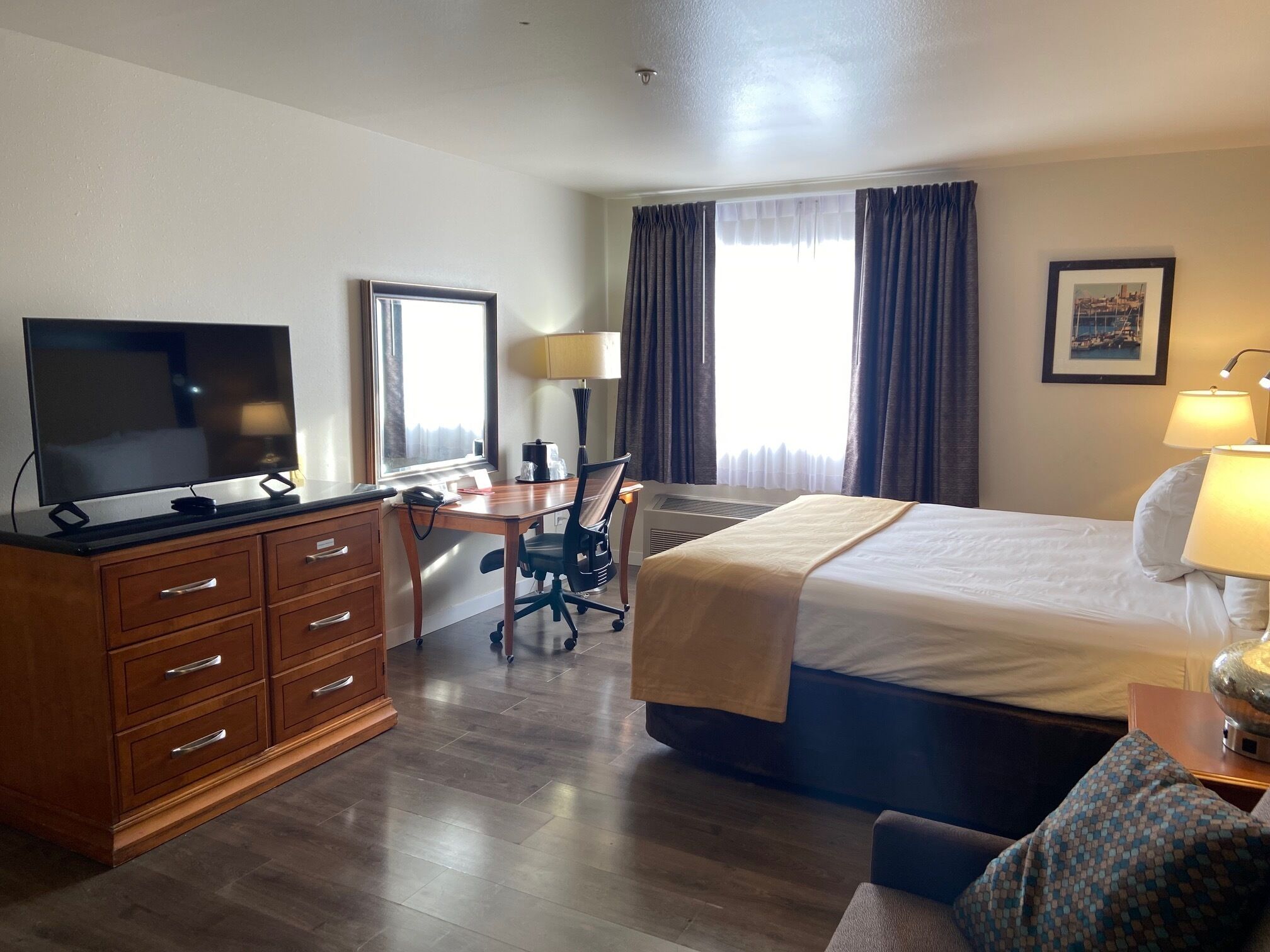 Une chambre d’hôtel comprenant un lit, un bureau avec une chaise, une télévision, une commode et une fenêtre avec des rideaux.