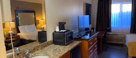 A hotel room with a bed, a desk with a computer, a coffee maker, a TV, and a bathroom with a sink and mirror.