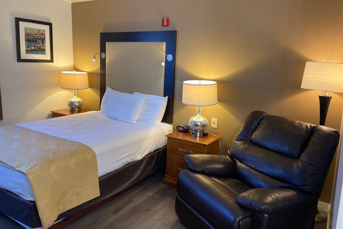 A hotel room with a bed, two bedside lamps, a black leather chair, and a wooden nightstand.