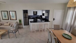 A modern living room with a sofa, a coffee table, and a dining area with a table and chairs. There is a kitchen in the background with a refrigerator and cabinets. - I 203, Stanza Mare, 2 bed, Private Beach, Bavaro , Punta Cana, 24 hrs security
 (Bavaro)