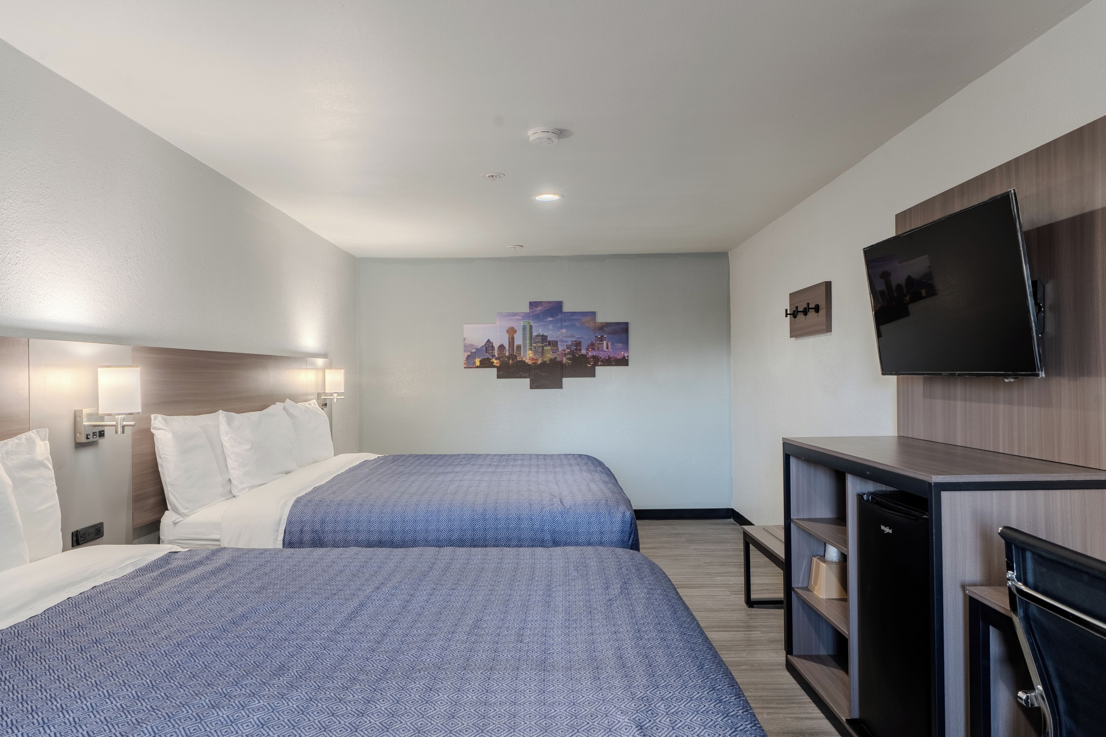 A hotel room with two beds, a flat-screen TV, a small refrigerator, and a cityscape artwork on the wall.