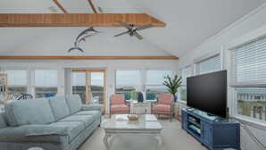 Un soggiorno ampio con soffitto alto, una ventola a soffitto, un'ampia televisione a schermo piatto, un divano chiaro, due poltrone rosa e un tavolino da caffè in vetro.