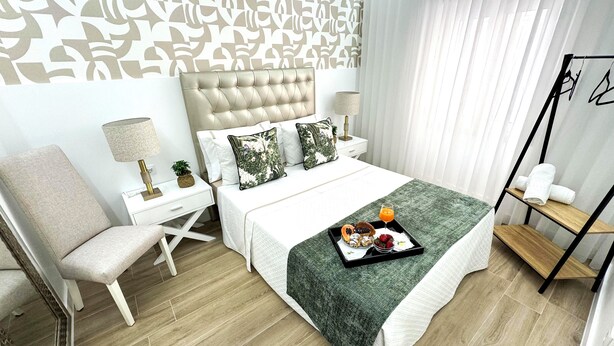 A modern hotel room with a tufted headboard, a bed with a green throw, a side table with a lamp, a chair, and a tray with breakfast items.