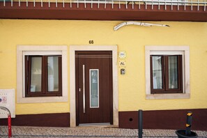 A building with a yellow facade, a brown door, and two windows with brown frames. - Apartamento Pérolo DO MAR (NAZARÉ)