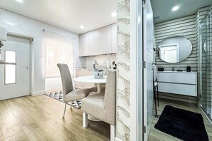A modern kitchen with a round island, a dining area with a chair, and a bathroom with a mirror and towel rack. - Apartamento Pérolo DO MAR (NAZARÉ)