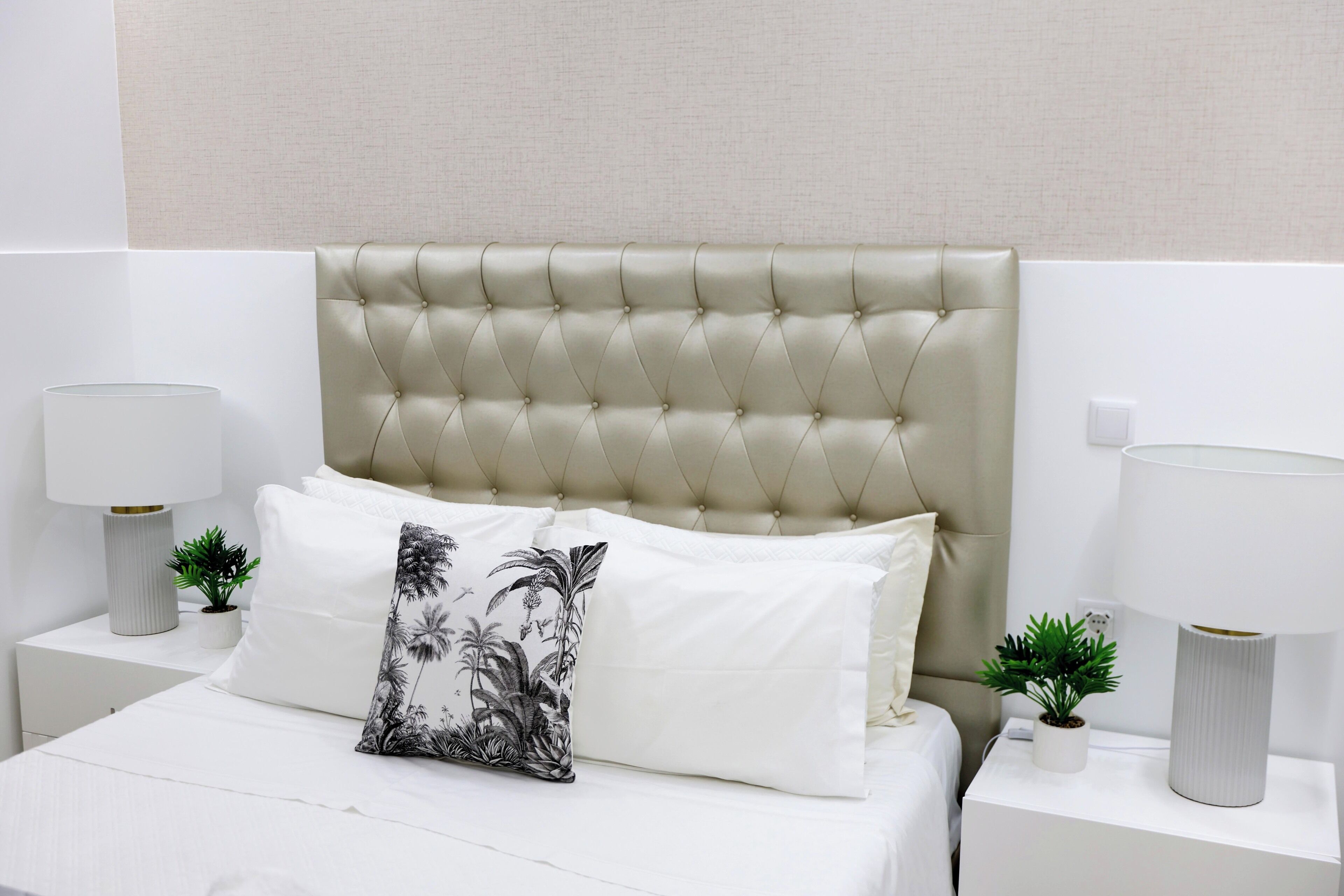 A neatly made bed with a tufted headboard, white bedding, and a decorative pillow. There are two bedside tables with lamps and small potted plants.