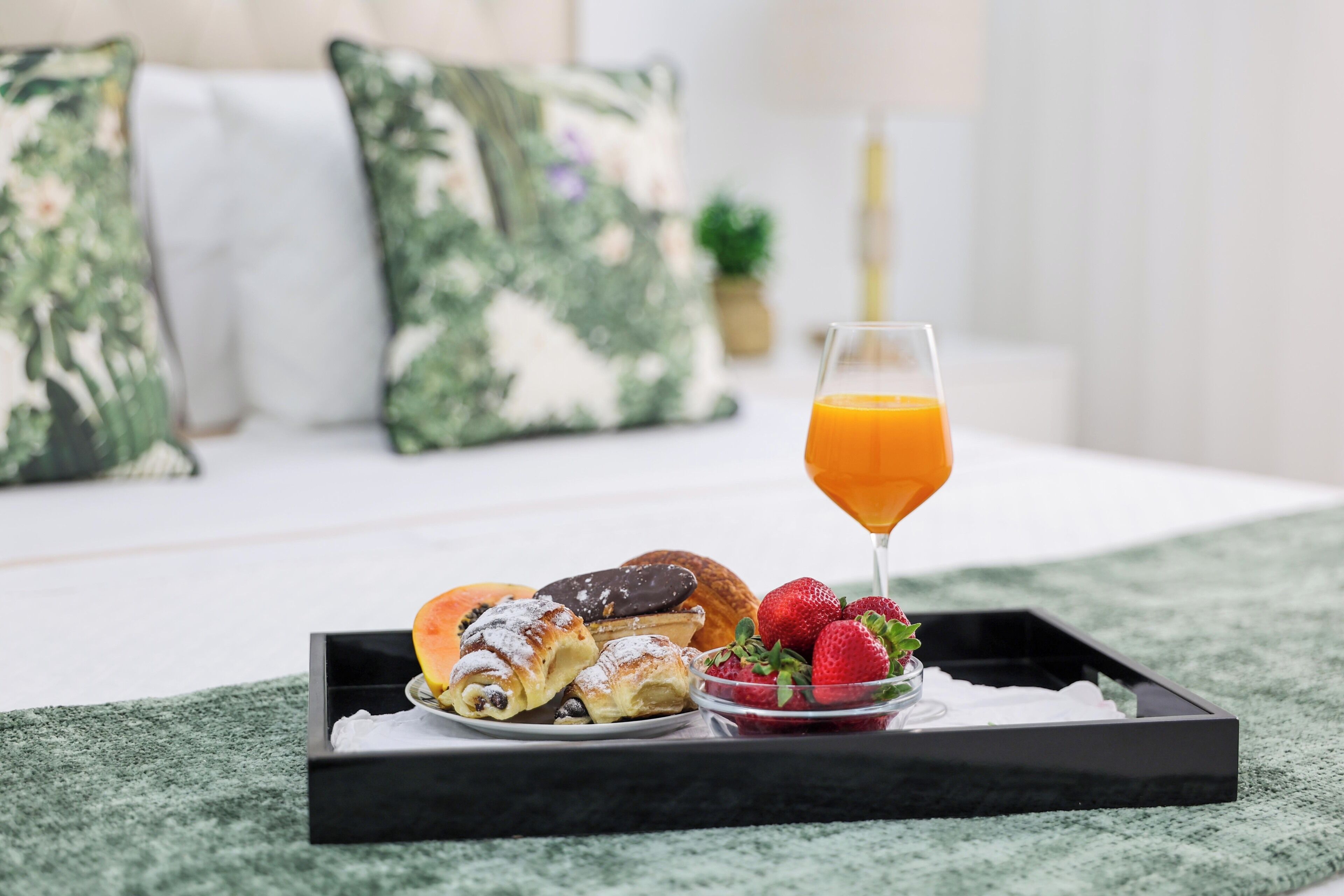 A breakfast tray with pastries and strawberries on a bed with a green blanket and floral pillows.