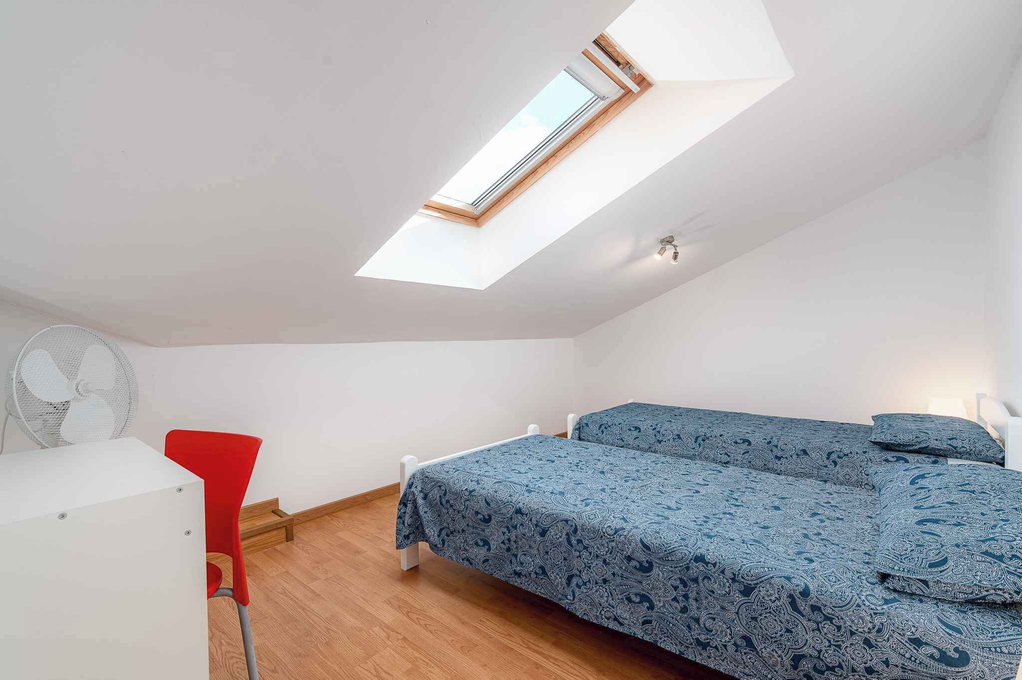 A small, bright room with a skylight, a bed with patterned bedding, a red chair, and a white desk.