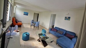 Un salon avec un canapé bleu, un fauteuil orange et une table basse en verre. On y trouve deux chaises et un pouf bleu.
