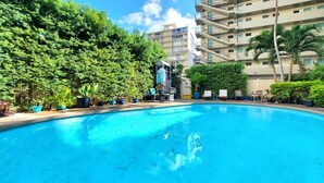 A swimming pool surrounded by lush greenery and a multi-story building with balconies. - Calm, Quiet, New, & Clean Waikiki Queen Bed Studio - WAIKIKI GRAND HOTEL® 804 (Honolulu)