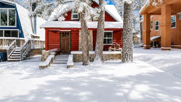 A snow-covered wooden cabin with a stone foundation, a red door, and a porch with outdoor seating.