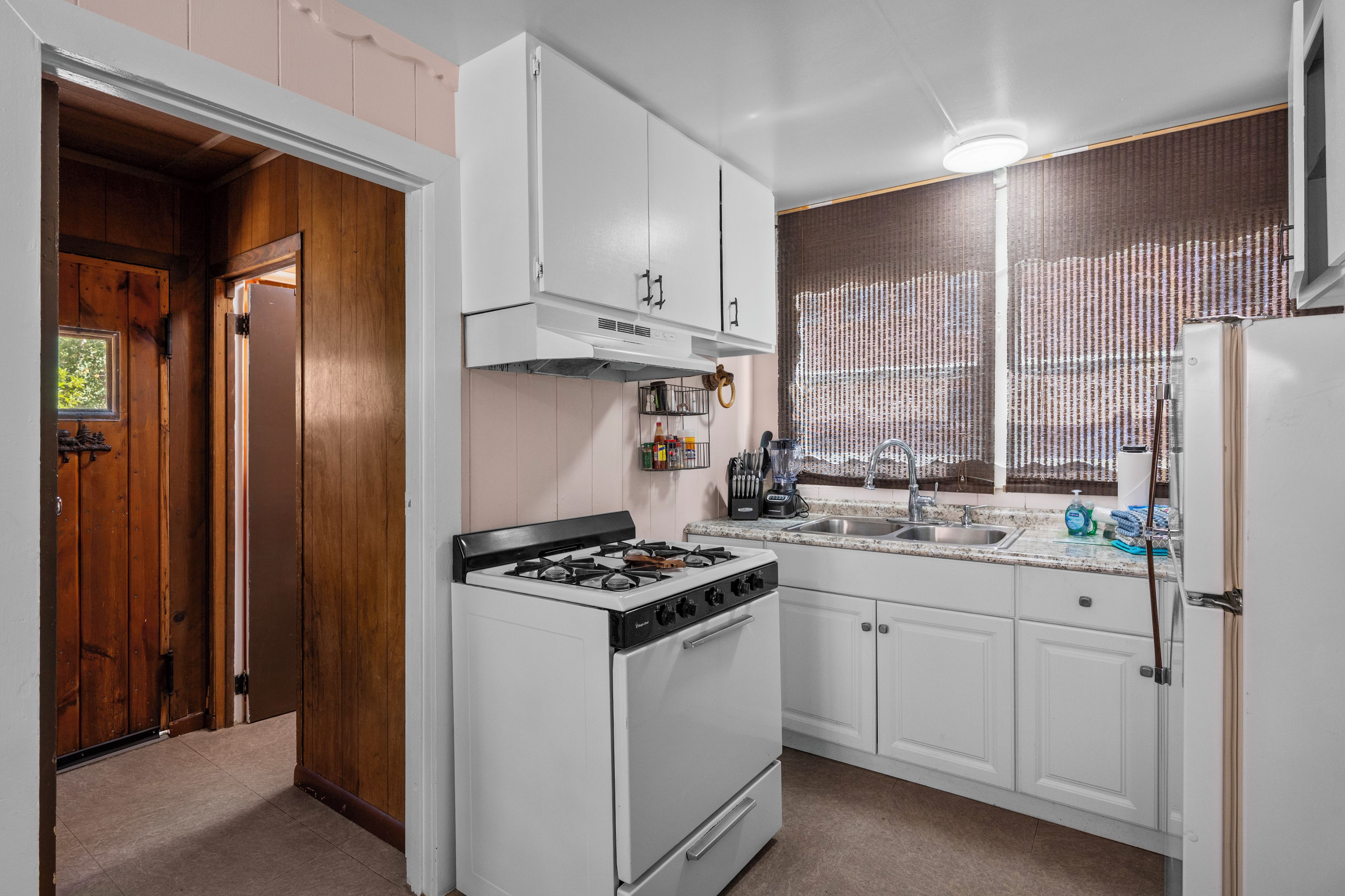 A kitchen with white cabinets, a stove, a refrigerator, and a window with blinds.