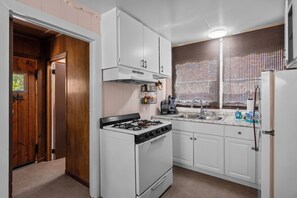 A kitchen with white cabinets, a stove, a refrigerator, and a window with blinds. - Lake Cottage (Big Bear Lake)