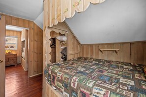 A bedroom with a sloped ceiling, wooden walls, a quilted bedspread, and a closet with hanging clothes. - Lake Cottage (Big Bear Lake)