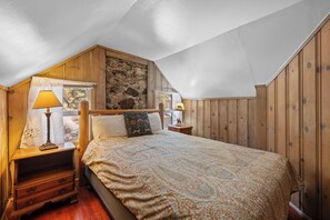 A bedroom with a sloped ceiling, wooden walls, a bed with patterned bedding, two bedside lamps, and a nightstand with a lamp. - Lake Cottage (Big Bear Lake)