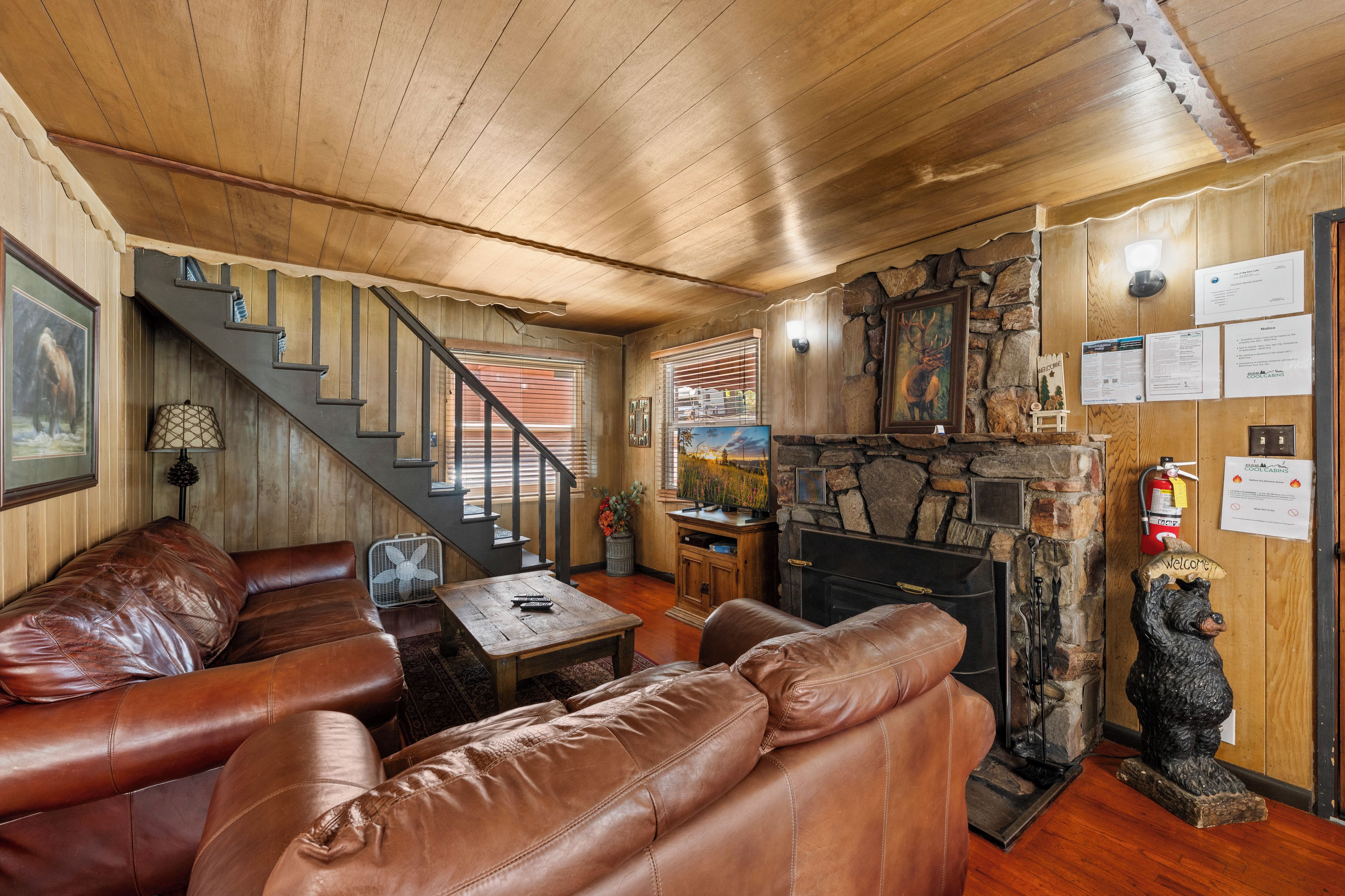 A cozy cabin interior with a stone fireplace, leather seating, a staircase, and a mounted deer head.