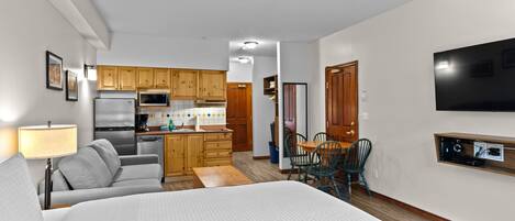 A modern hotel room with a large bed, a sofa, a dining table, and a kitchen area with wooden cabinets and appliances.