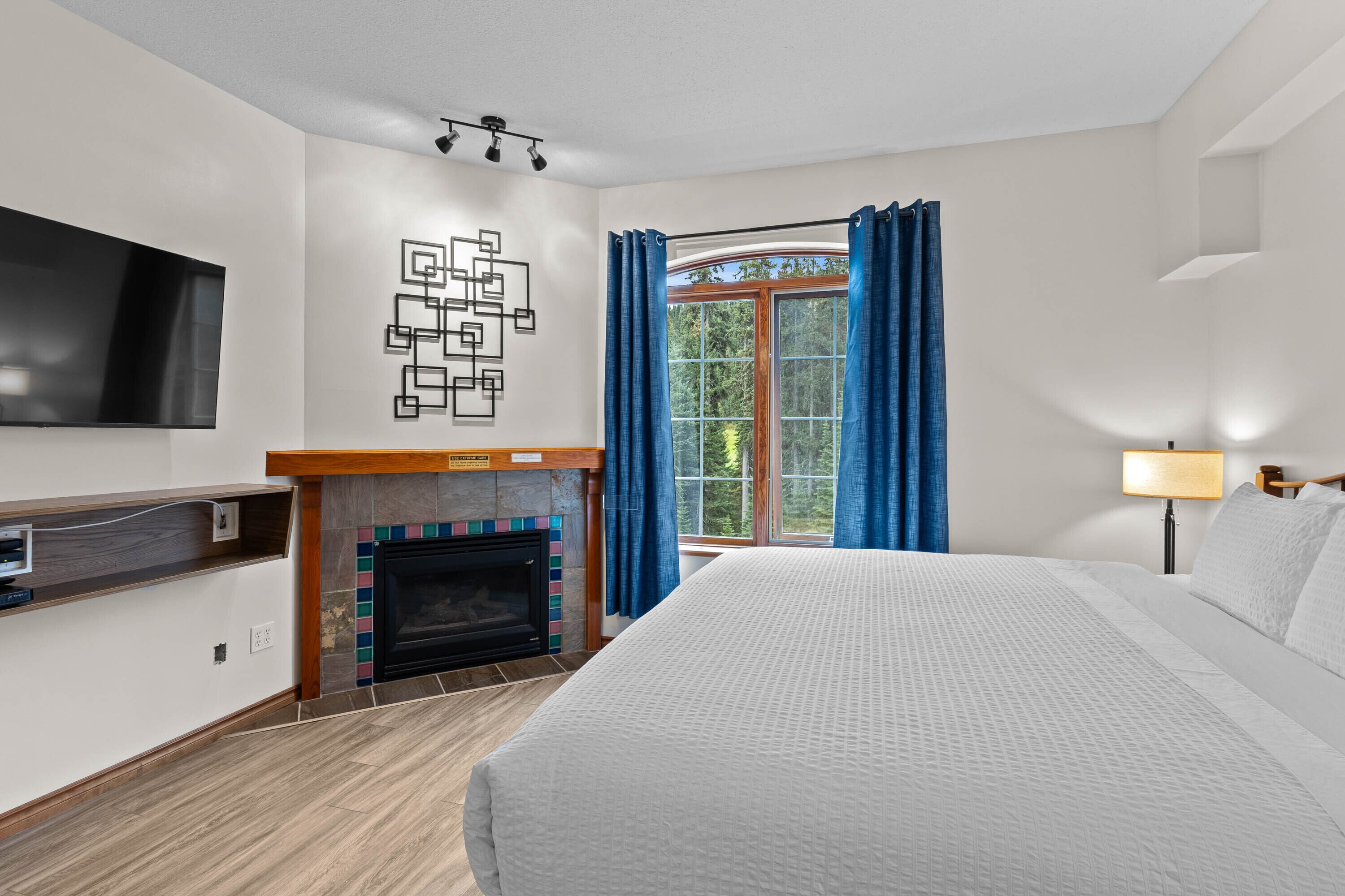 A modern hotel room with a large bed, a fireplace, a flat-screen TV, and a window with curtains.