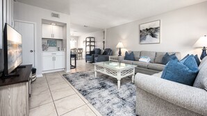 A modern living room with a sofa, coffee table, and TV. There is a kitchen area with cabinets and a dining table. - Gulf Winds East 22 * Fall Specials * Across from beach * Salt Water Vacations (Destin)