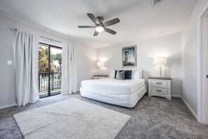 A bedroom with a bed, a ceiling fan, two bedside tables with lamps, a painting on the wall, and a view through a glass door.
