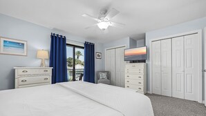 Une chambre à coucher avec un lit, un ventilateur de plafond, une télévision, une armoire et une fenêtre avec des rideaux.
