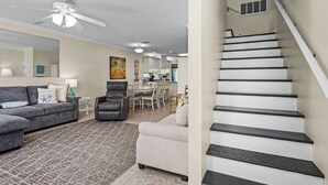 Un salon moderne avec un canapé gris, un fauteuil blanc et un tapis à motifs. On y trouve un escalier aux marches sombres menant à un niveau supérieur.