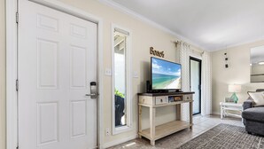 Un salon avec un téléviseur posé sur un meuble en bois, une porte donnant sur l’extérieur et un panneau fixé au mur sur lequel on peut lire « Plage ».